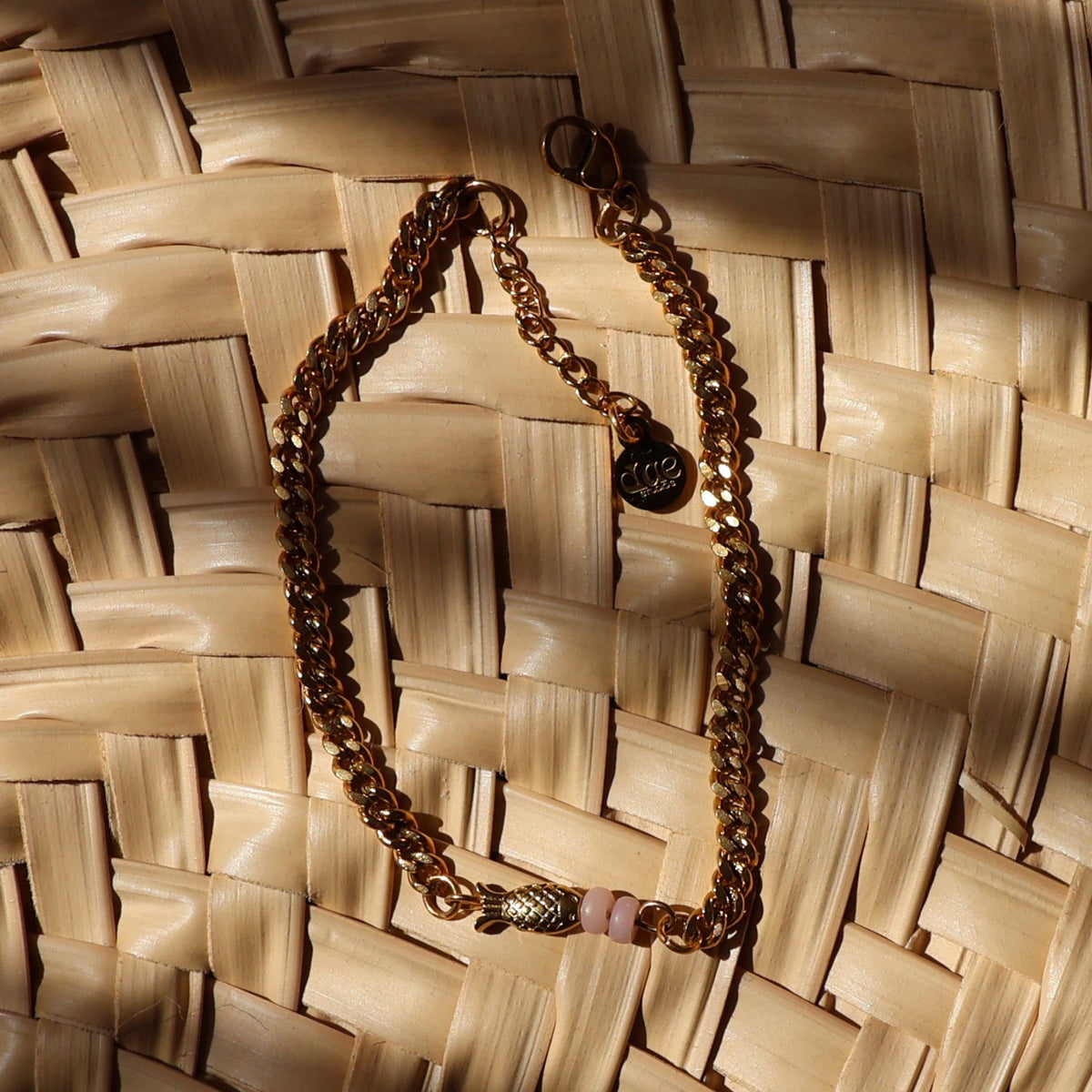 A gold chain necklace with small pink accent beads and gold-plated fish bead, displayed on a woven straw mat under warm, direct sunlight.