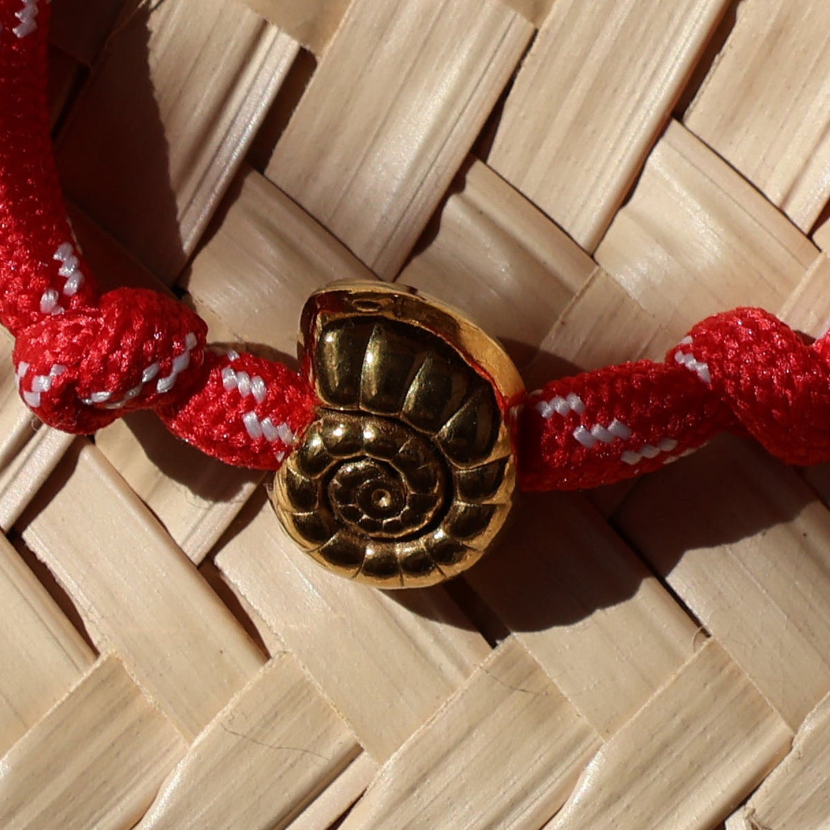 A red braided cord bracelet with gold-toned metal charms, including a round medallion and a small dangling tag, displayed on a woven straw hat under warm, directional sunlight.