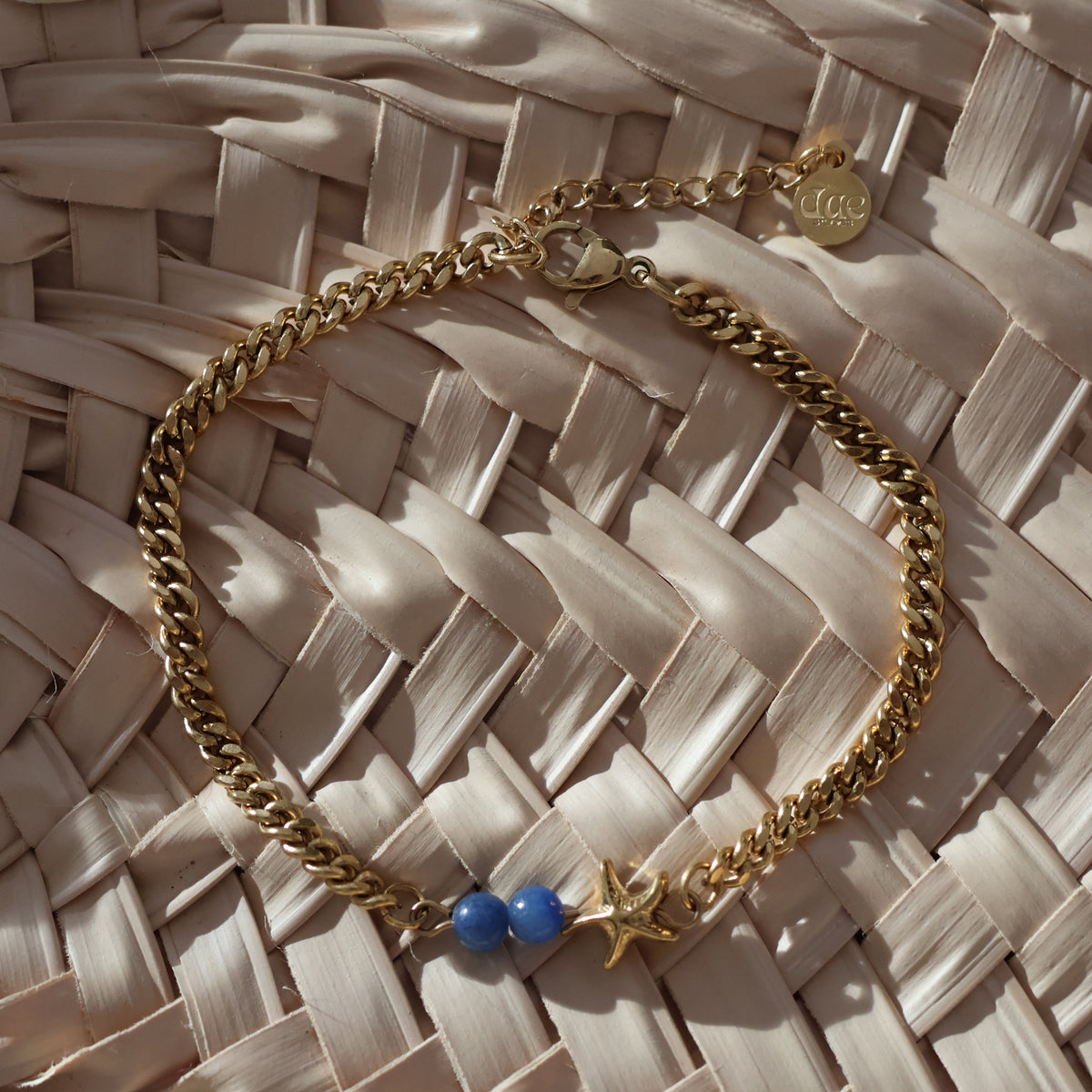 A gold chain bracelet with a star charm, two blue beads, and a small round charm, displayed on a woven straw mat.