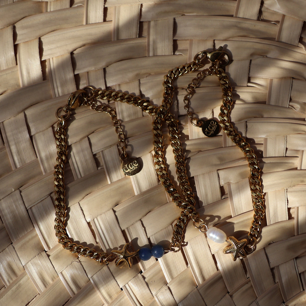 Two gold-toned chain bracelets with small charms, one featuring a blue bead and the other a pearl, displayed on a woven straw mat.