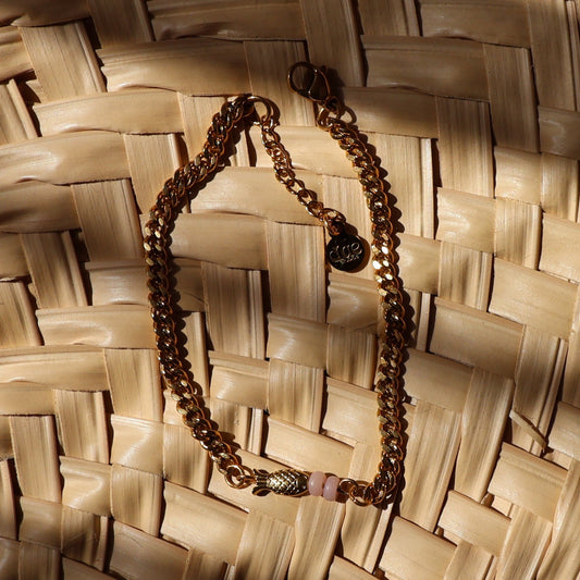A gold chain necklace with small pink accent beads and gold-plated fish bead, displayed on a woven straw mat under warm, direct sunlight.