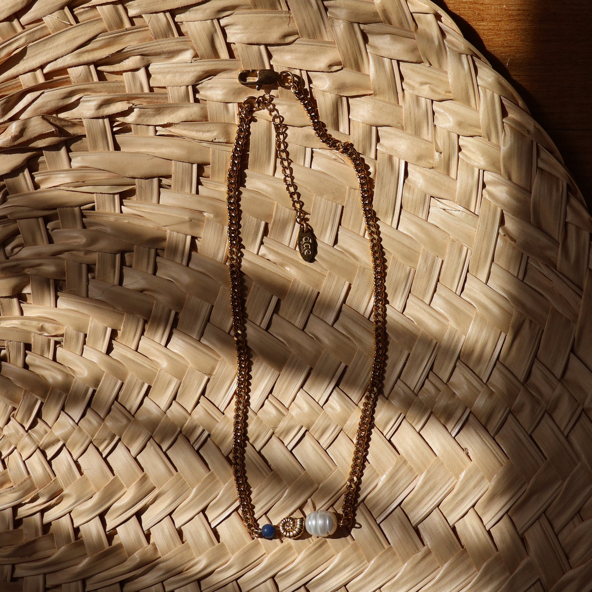 A close-up of a gold chain necklace with pearl and beads, laid on a woven straw background, with dappled shadows.
