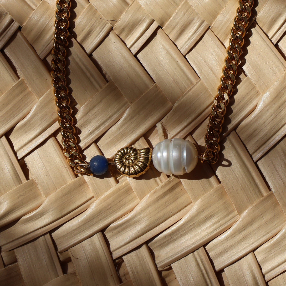 A gold chain necklace featuring a central gold-toned nautilus shell charm, a white pearl, and a small blue adventuring bead, displayed on a woven background under dappled sunlight.