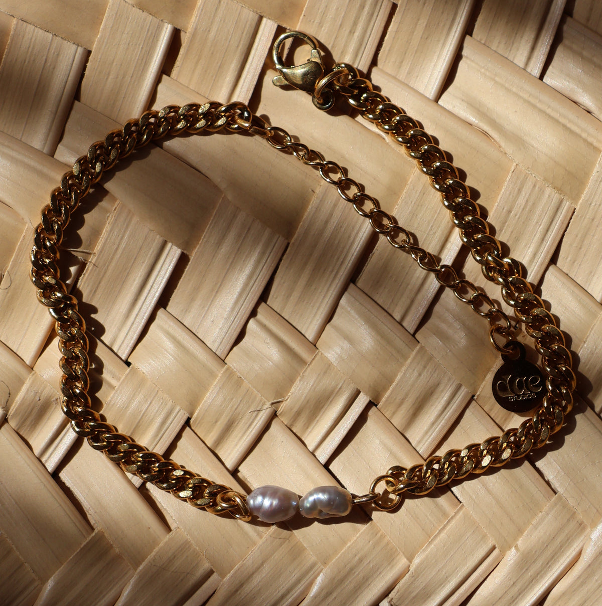 A gold plated chain necklace with two grey pearl accents, displayed on a woven mat.