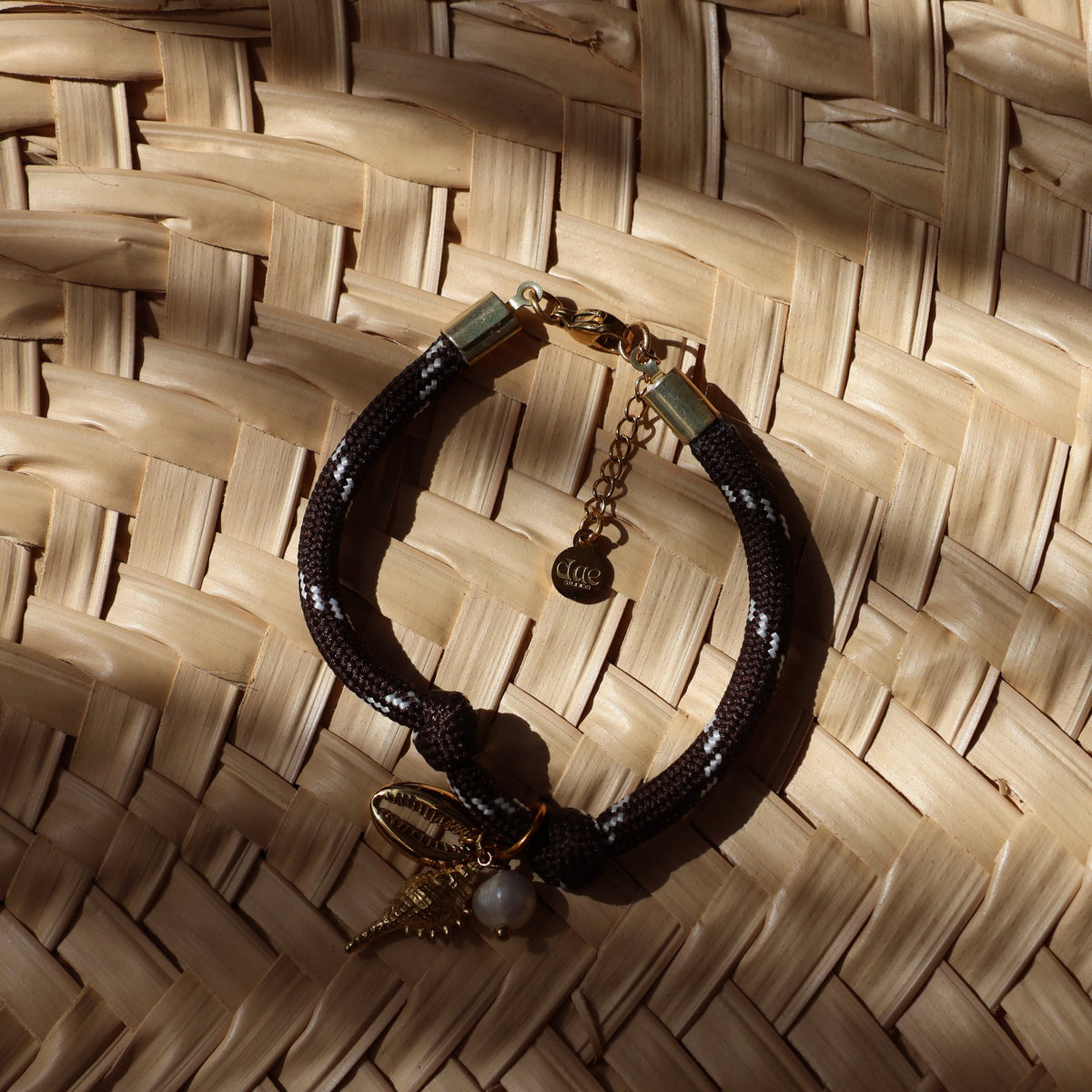 A black braided cord bracelet with gold-toned hardware, a small white pearl bead, and a gold-toned pendant, displayed on a woven straw background.