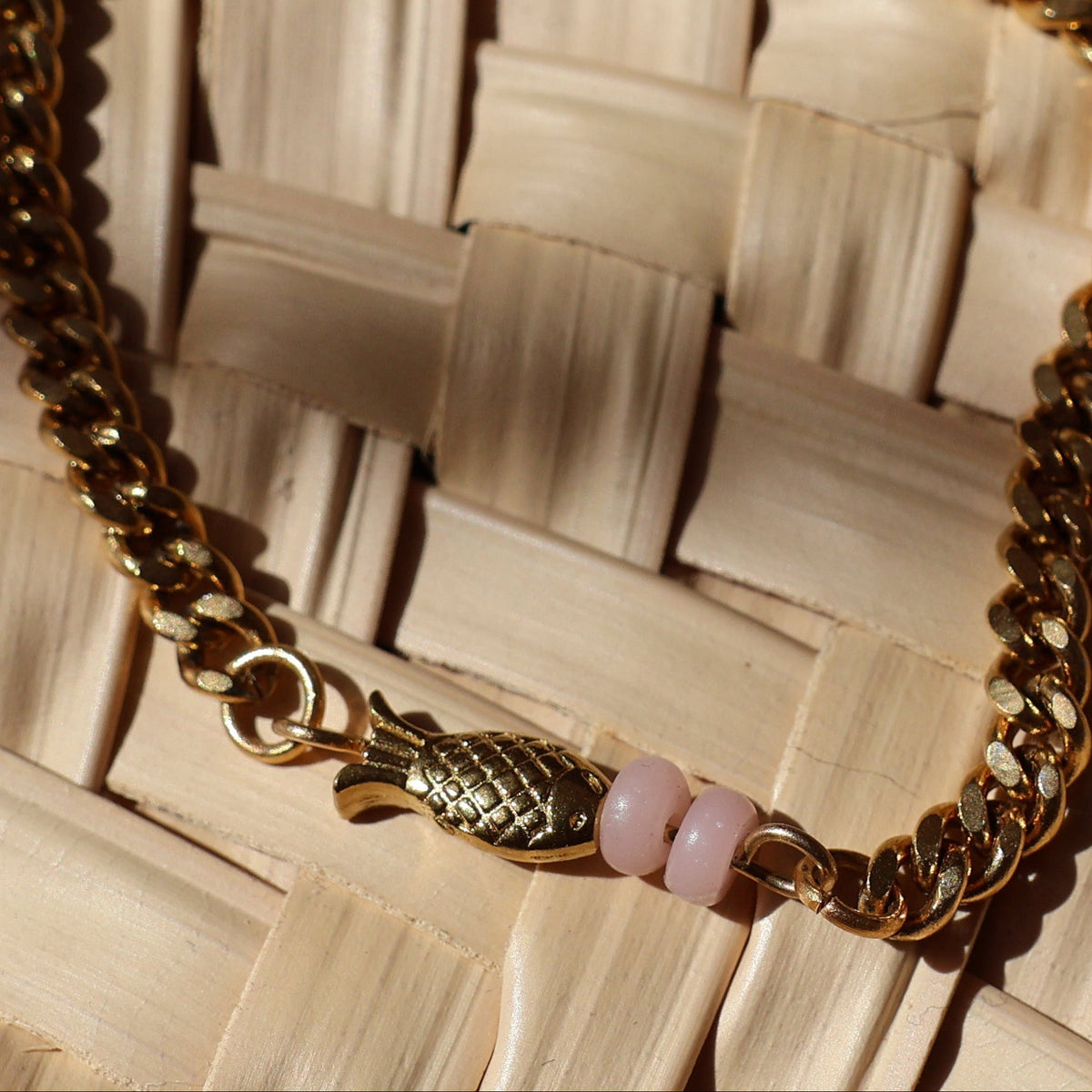 A gold chain necklace with small pink accent beads and gold-plated fish bead, displayed on a woven straw mat under warm, direct sunlight.
