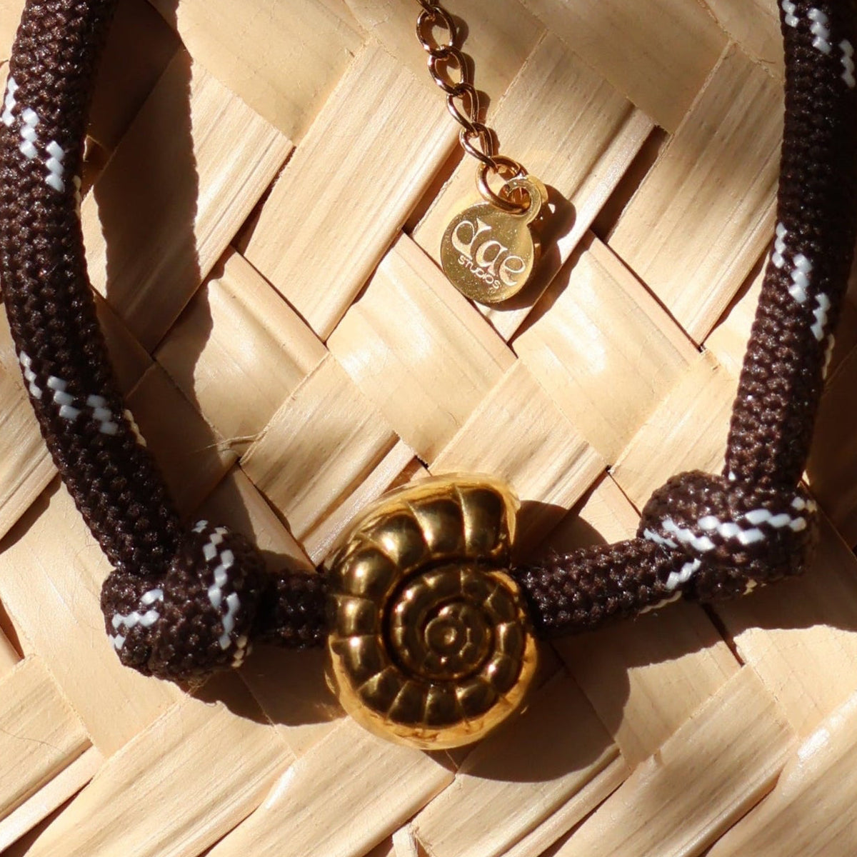 A dark brown leather bracelet with gold-toned hardware and a nautilus gold-plated charm, displayed on a straw mat with dappled light.