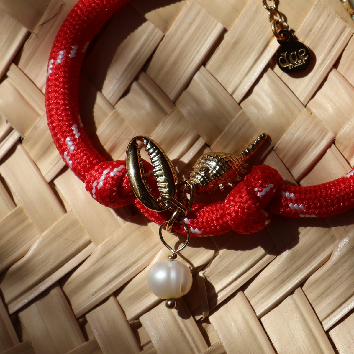 A red braided cord bracelet with white woven accents, featuring a gold-tone conch shell charm, a white pearl bead, and a small gold tag, displayed on a light wood woven mat.