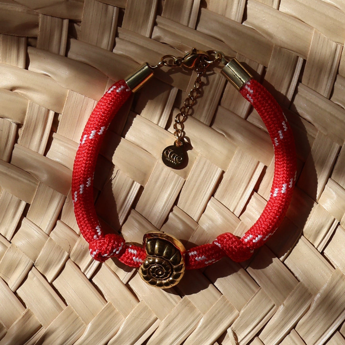 A red woven bracelet with a gold-toned charm resting on a large woven straw hat, under warm natural sunlight with soft shadows.